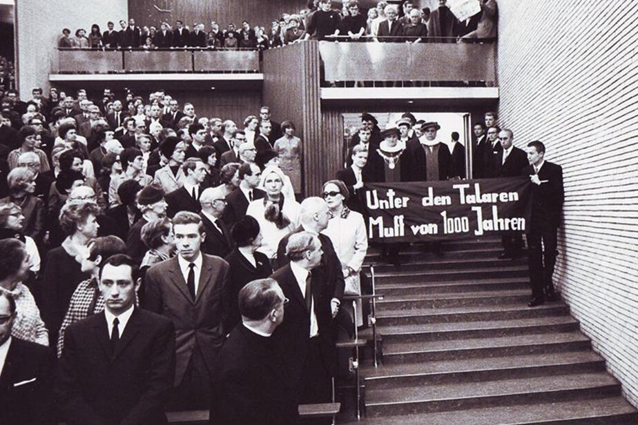 Standing room only! Audimax on 9 November 1967. Two students carry a banner bearing the words: “Under these robes—1,000 years of rot!”