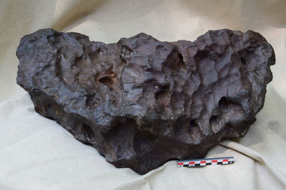 The photo shows a black meteorite against a beige background. In front of the meteorite is a linear measure in black white and red. The surface of the meteorite has a lot of structure.