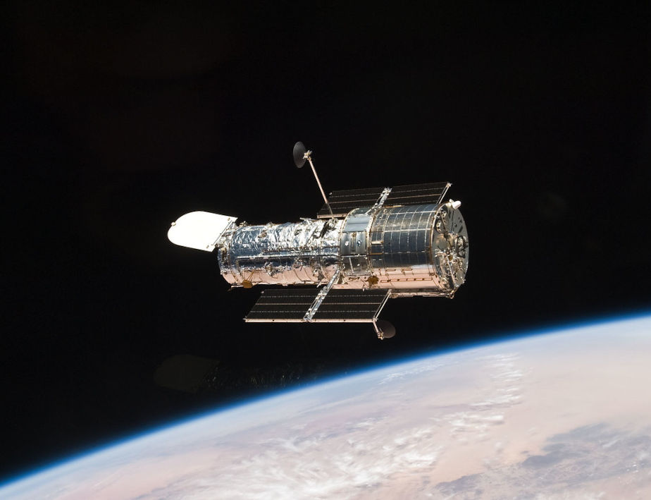 Die farbige Abbildung zeigt in der Mitte silbrig glänzend das im Weltraum schwebende Hubble-Teleskop. Unten ist hellblau die Erde und Erdatmosphäre zu sehen.