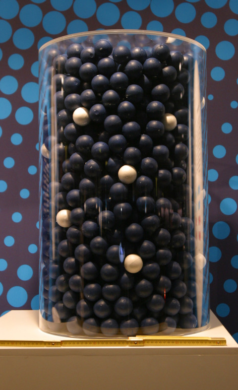 Die farbige Abbildung zeigt ein zylinderförmiges Gefäß, das mit vielen schwarzen und einigen weißen Kugeln gefüllt ist. Das Gefäß steht auf einem beigen Sockel und vor einer schwarzen Wand mit blauen Kreisen.