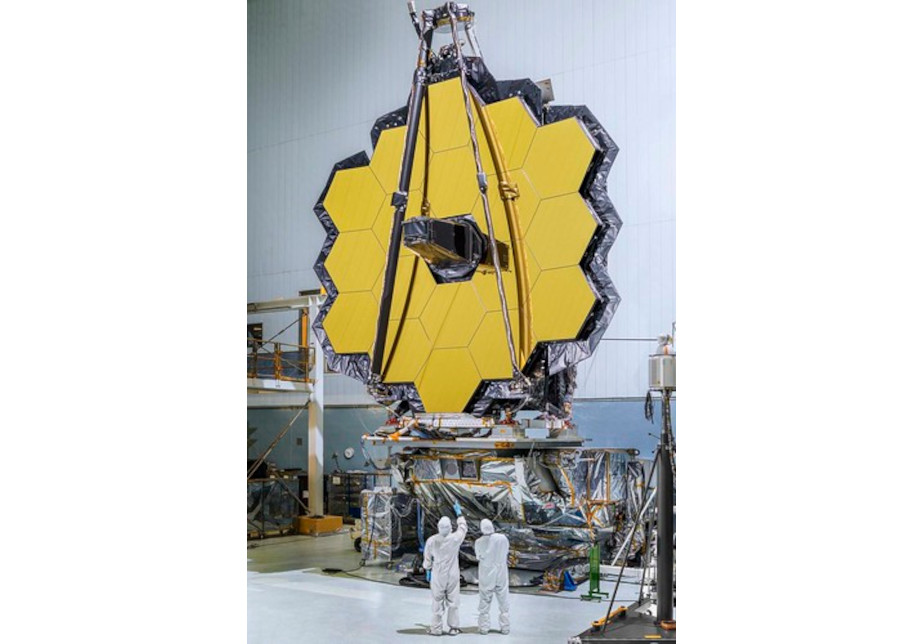 Die farbige Abbildung zeigt den Spiegel des James-Webb-Teleskops in einer großen Halle. Zwei Menschen in weißen Schutzanzügen stehen davor.