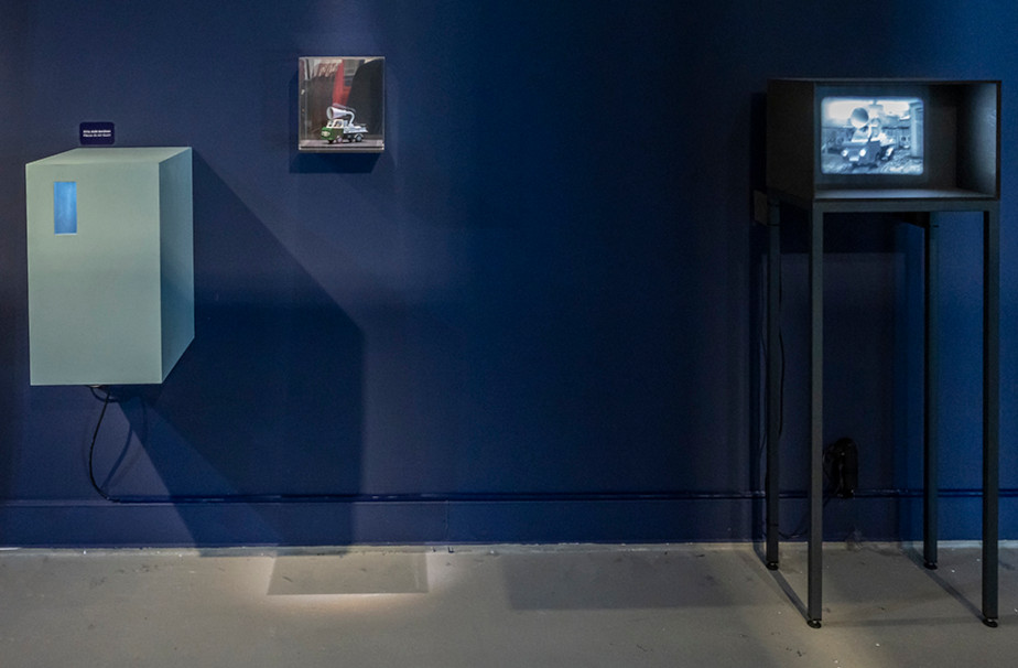 In front of a dark blue wall we can see three objects: there is a white box hanging on the far left with a small hole to look into. Next to it is a small glass case containing a model car with a large funnel on the roof. On the right there is an old-fashioned TV that is currently showing a black and white picture of the same car with a funnel on the roof.