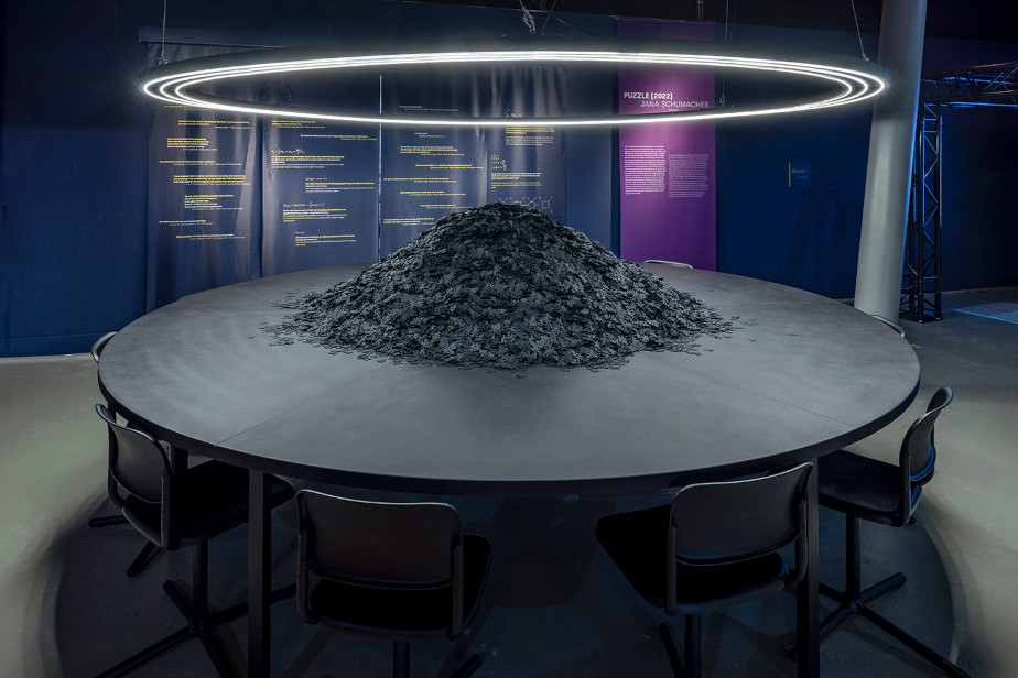 Zu sehen ist ein großer Konferenztisch, um den herum einige Stühle stehen. Von oben wird der Tisch von einer ringförmigen Leuchtstoffröhre beleuchtet. Auf dem Tisch türmen sich tausende von schwarzen Puzzleteilen.