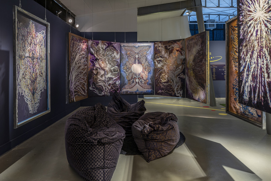 The photo from the exhibition shows abstract pictures in metal frames that are suspended from the ceiling in a kind of semicircle in front of the viewer. In between the picture frames, three dark brown beanbags lie on the floor. The colors of the pictures and their arrangement give the impression of stained glass windows in a church.