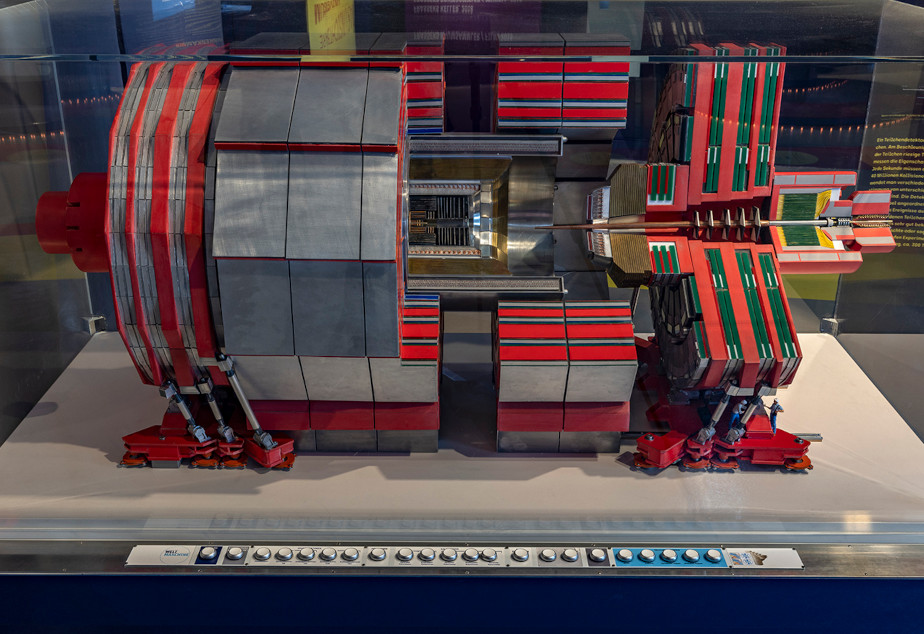In a display case there is a red and silver model of the CMS detector, which has the shape of a cut open cylinder. There is a row of buttons in front of the display case.