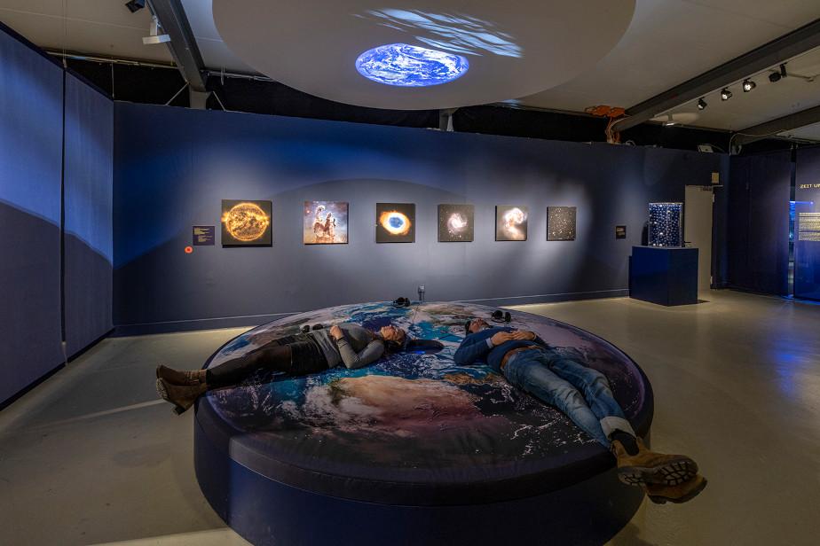 In the foreground, the photo shows a circular lounging area on which two people are lying with headphones and looking upwards. Above them hangs a screen, also circular, which currently shows an image of the Earth above the lunar horizon. In the background, images from the Hubble Space Telescope are hanging on a blue wall.