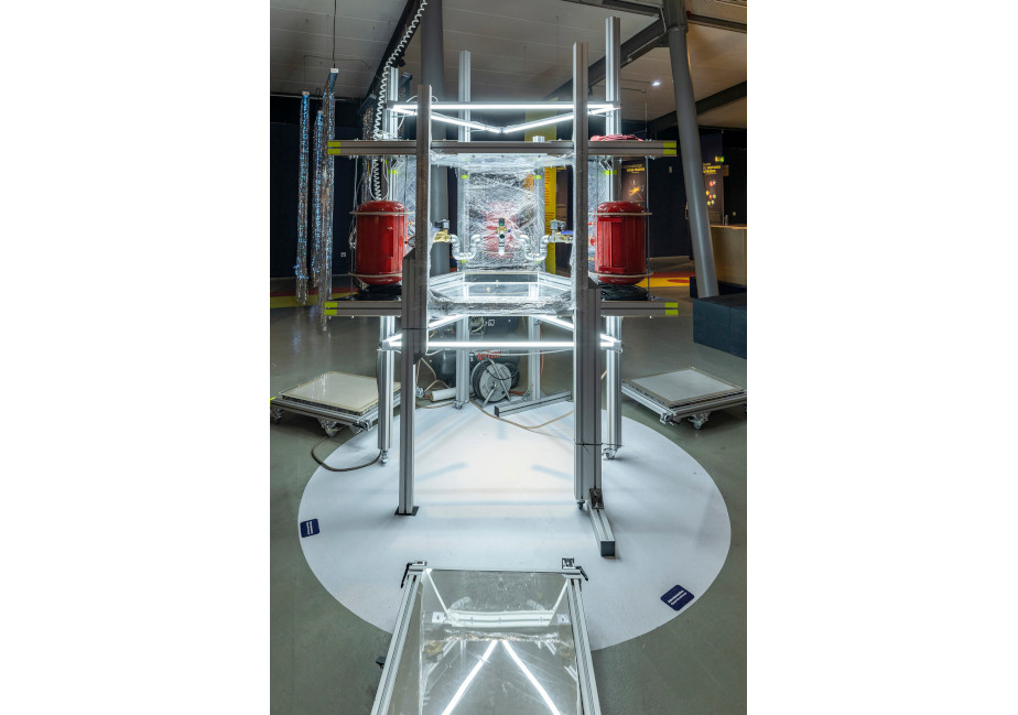 The photo from the exhibition shows a construction made of metal struts. To the left and right are red containers that look somewhat like fire extinguishers. On the floor around the construction are three litho stones.