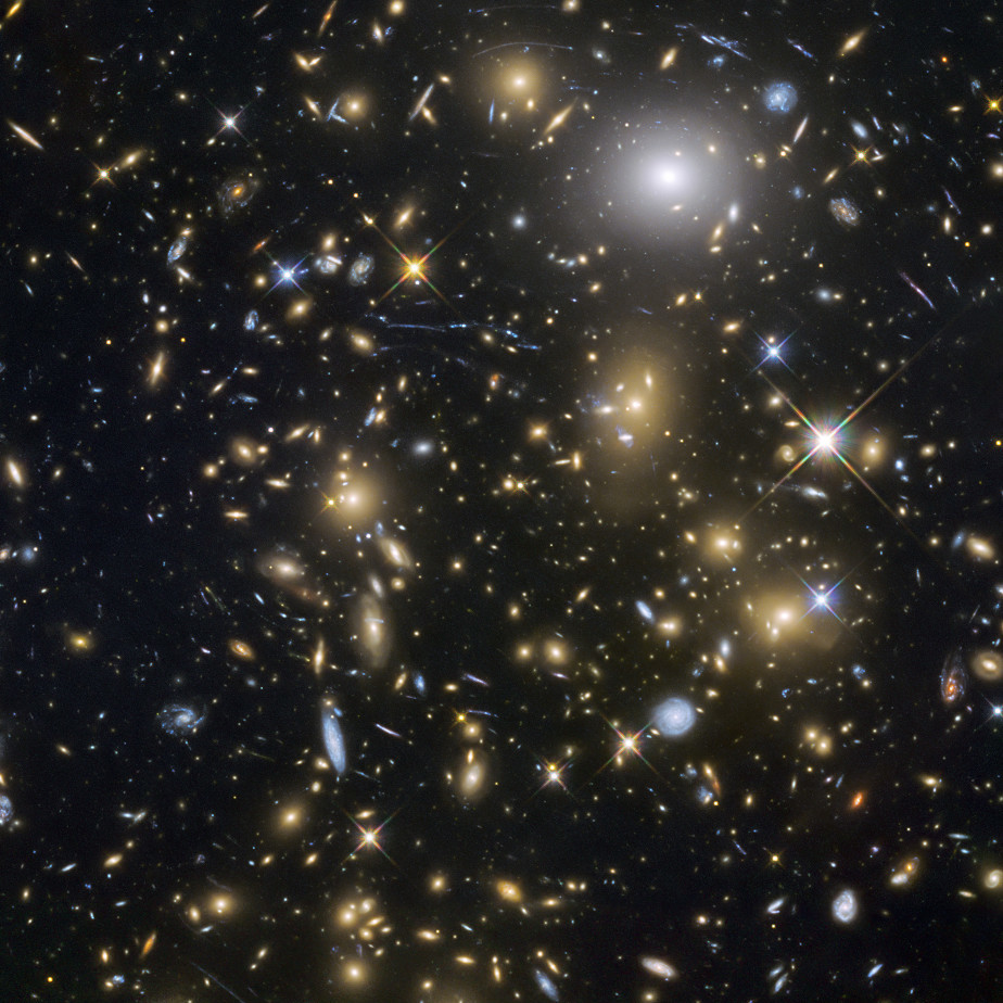 Die farbige Abbildung zeigt die Weiten unseres Universums. Vor schwarzem Hintergrund sind viele leuchtende Flecken in gelb, blau und weiß zu sehen, die teils wie Sterne, teils wie Spiralgalaxien aussehen.